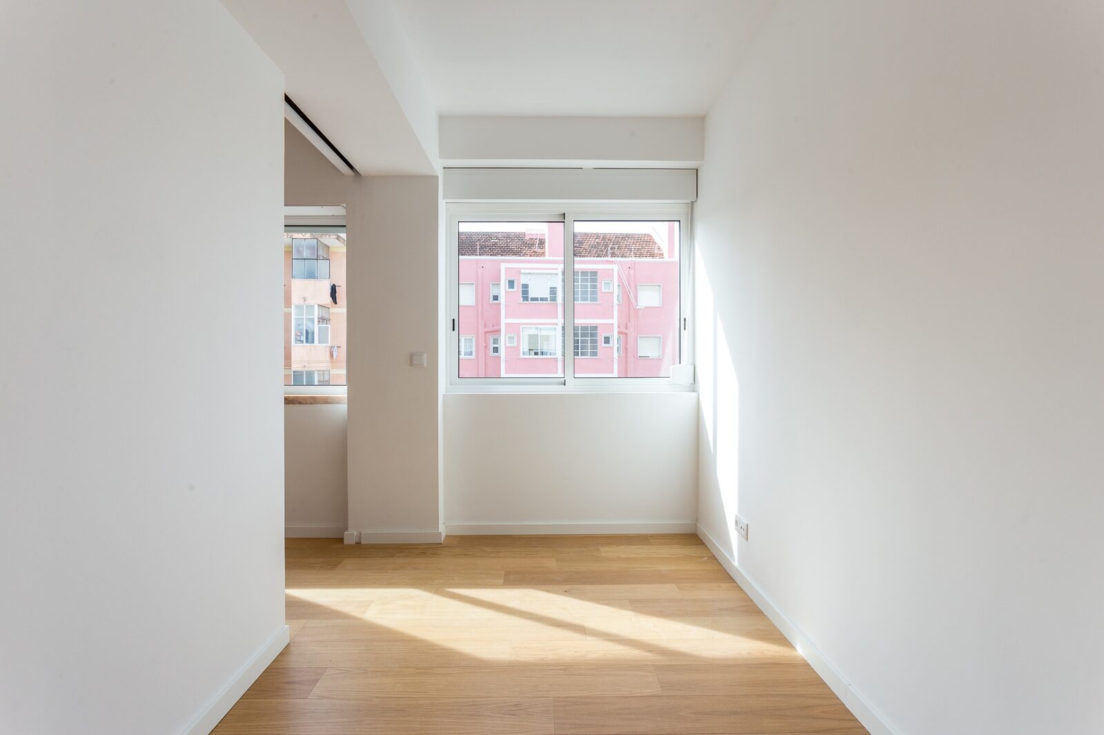 Quarto com vista depois — paredes brancas, pavimento em carvalho e janela com vista para edifício rosa típico da Barceloneta