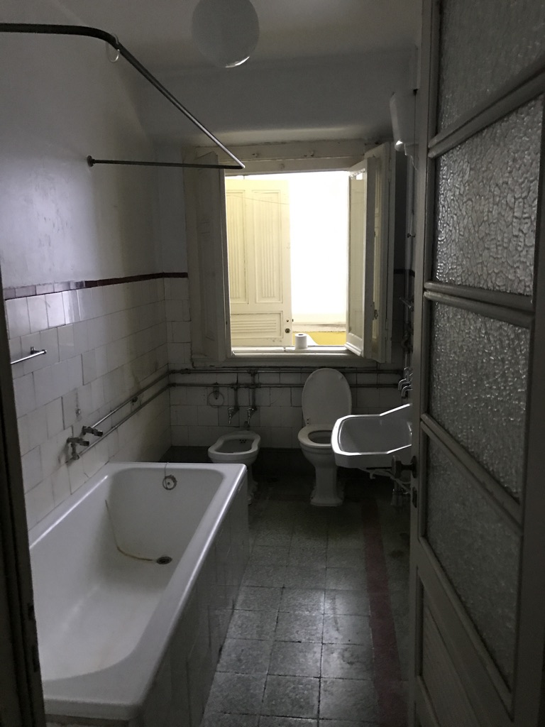 Casa-de-banho antiga com banheira em ferro, sanita, bidé, azulejos brancos com friso vermelho e janela ao fundo, Cabo Verde