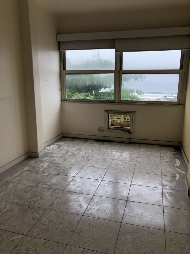 Quarto antes — paredes amareladas, pavimento em ladrilho com vestígios de tinta e janela com vista para o mar de Copacabana